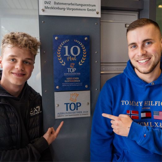 Zwei Azubis zeigen auf die Plakette "TOP-Ausbildungsbetrieb 2024"