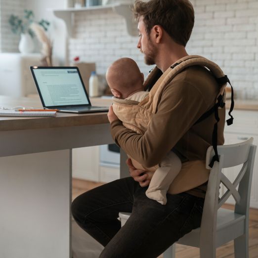 Foto eines Mannes mit einem Baby in einer Trage vor einem Laptop sitzend
