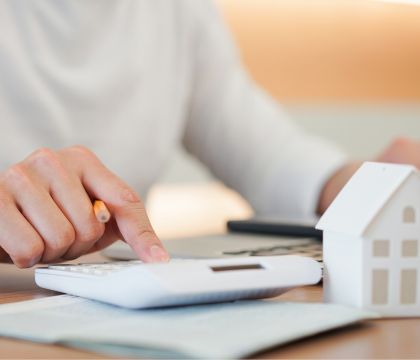 Bild: Ein Mann sitzt mit einem Taschenrechner an einem Schreibtisch vor einem Laptop. Vor dem Taschenrechner und dem Laptop steht die Miniatur eines Hauses.