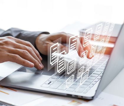 1)	Bild: Eine Person schreibt mit der einer Hand auf einem Laptop und nutzt mit der anderen das Touchpad. Über dem Laptop schweben mehrere Symbole von Dokumenten.