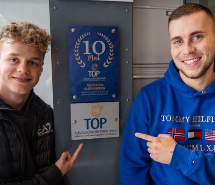 Zwei Azubis zeigen auf die Plakette "TOP-Ausbildungsbetrieb 2024"